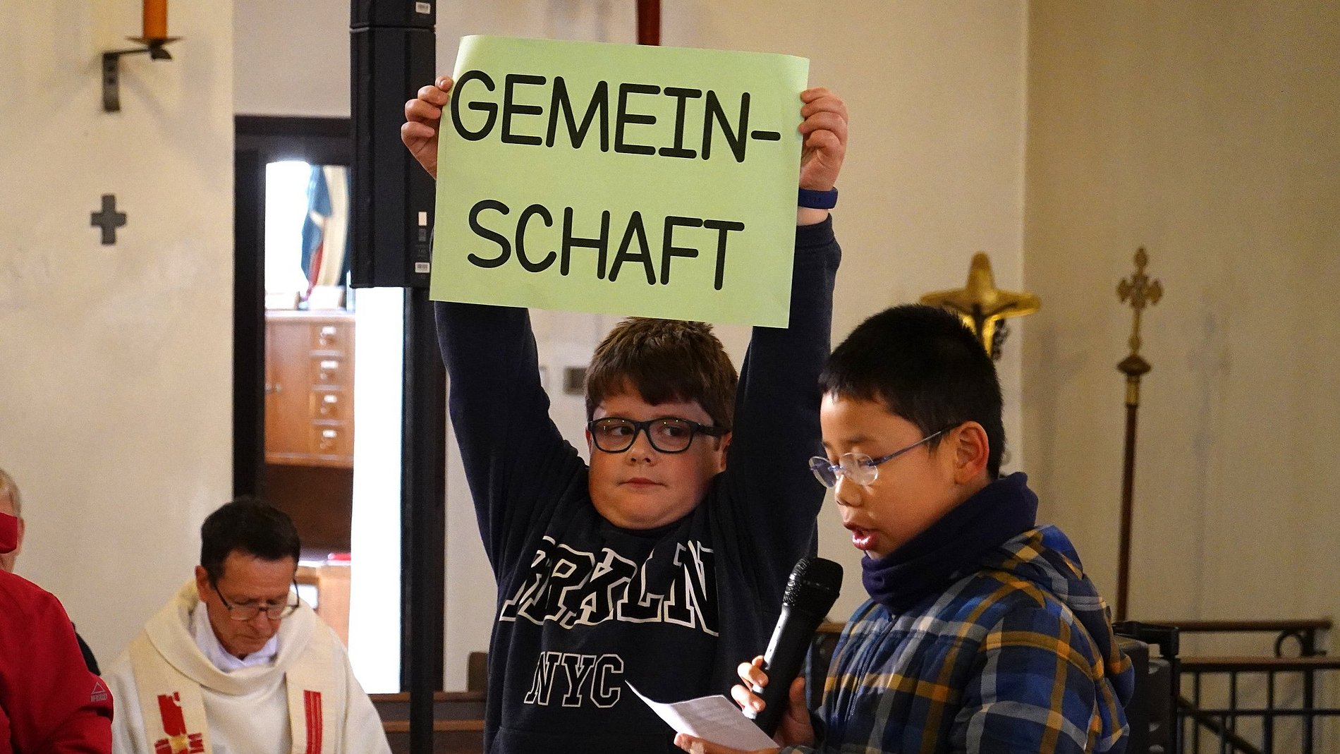 Ein Junge ließt eine Fürbitte, ein anderer hält ein Schild mit "Gemeinschaft" über seinem Kopf